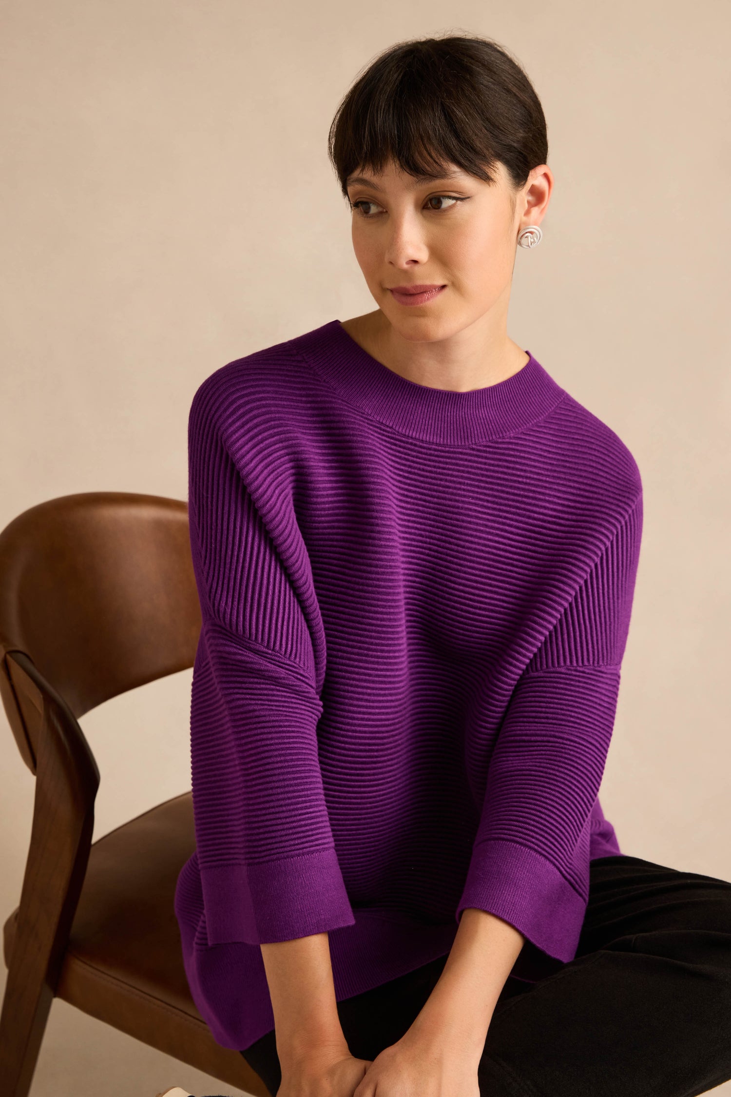 A person with short dark hair sits on a brown chair, looking slightly to the side against a neutral background, wearing the Wide Sleeve Purple Ribbed Knit.