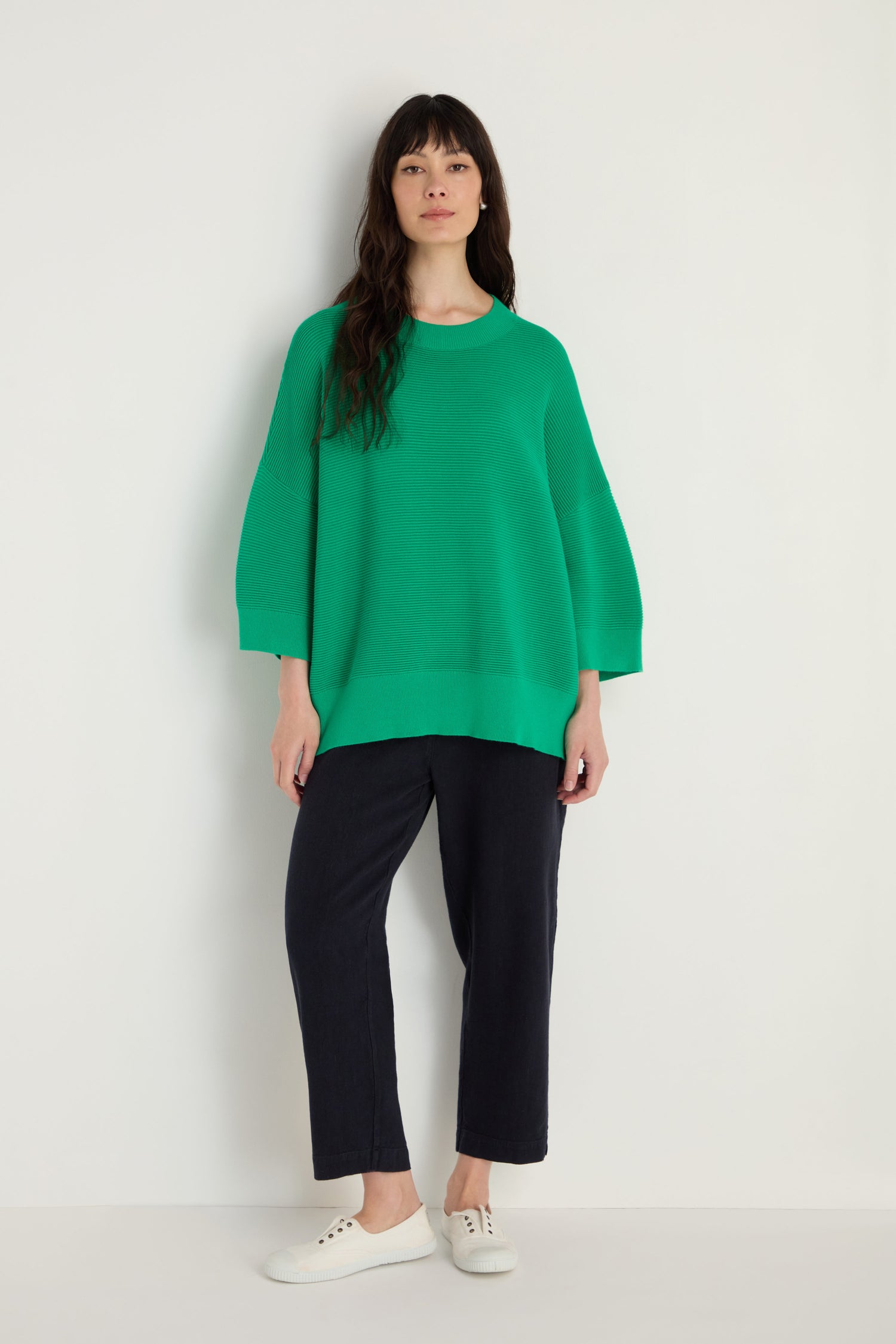 A woman stands against a white wall wearing the Wide Sleeve Ribbed Top in green, styled with black pants and white sneakers.