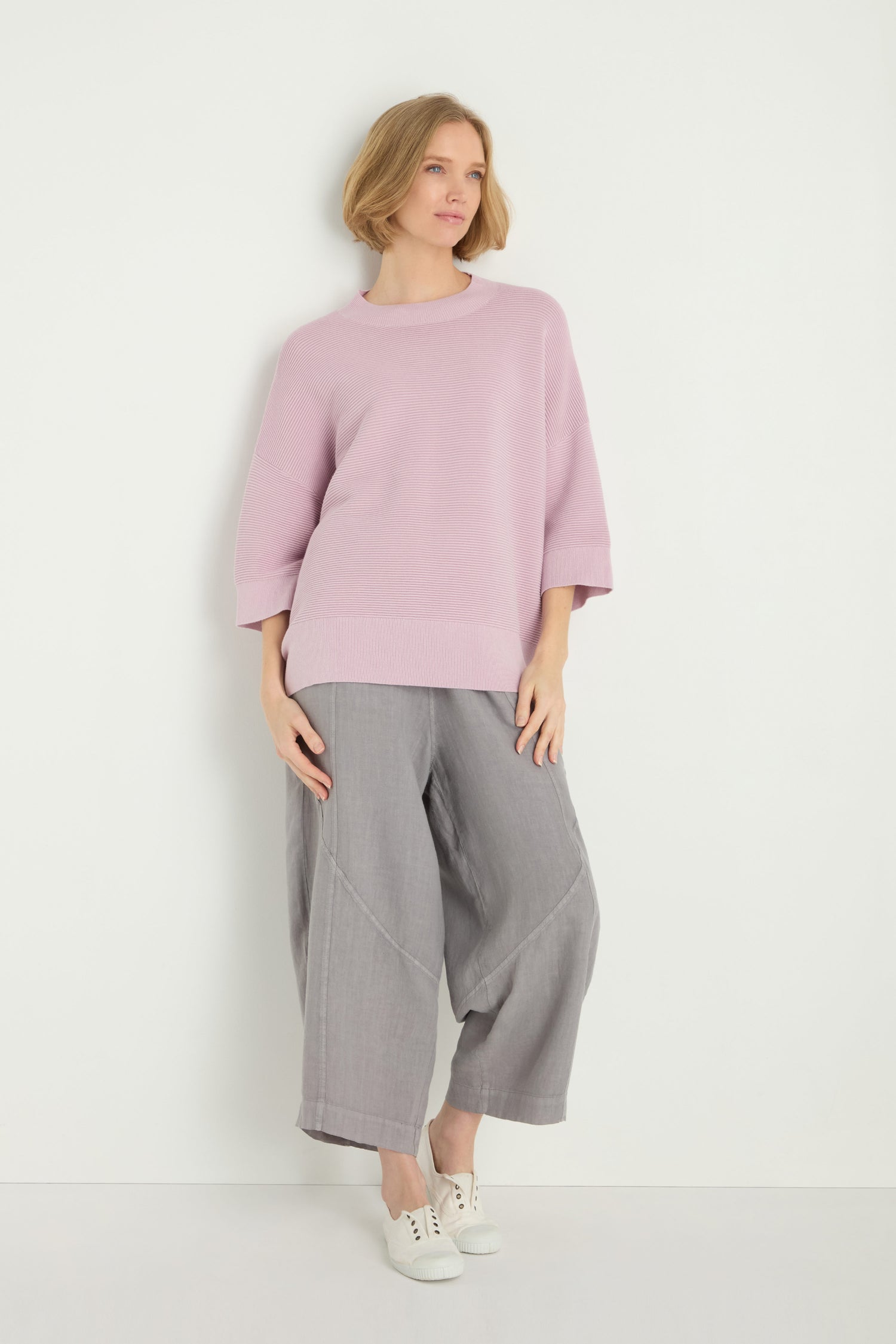 A person stands against a white wall wearing the Wide Sleeve Ribbed Top in light pink, featuring a boxy silhouette, styled with wide-leg gray pants and white sneakers.