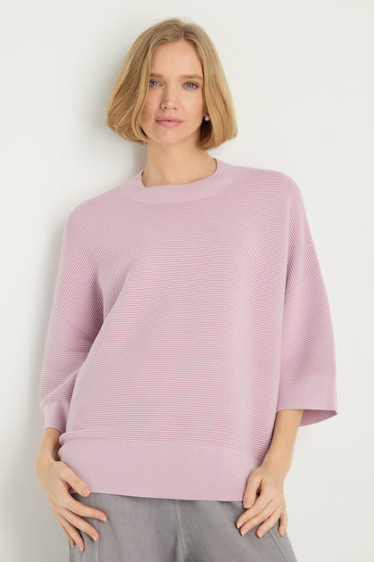 A woman with short blonde hair wears the Wide Sleeve Ribbed Top in light lavender with a ribbed texture and gray pants, standing against a plain white background.