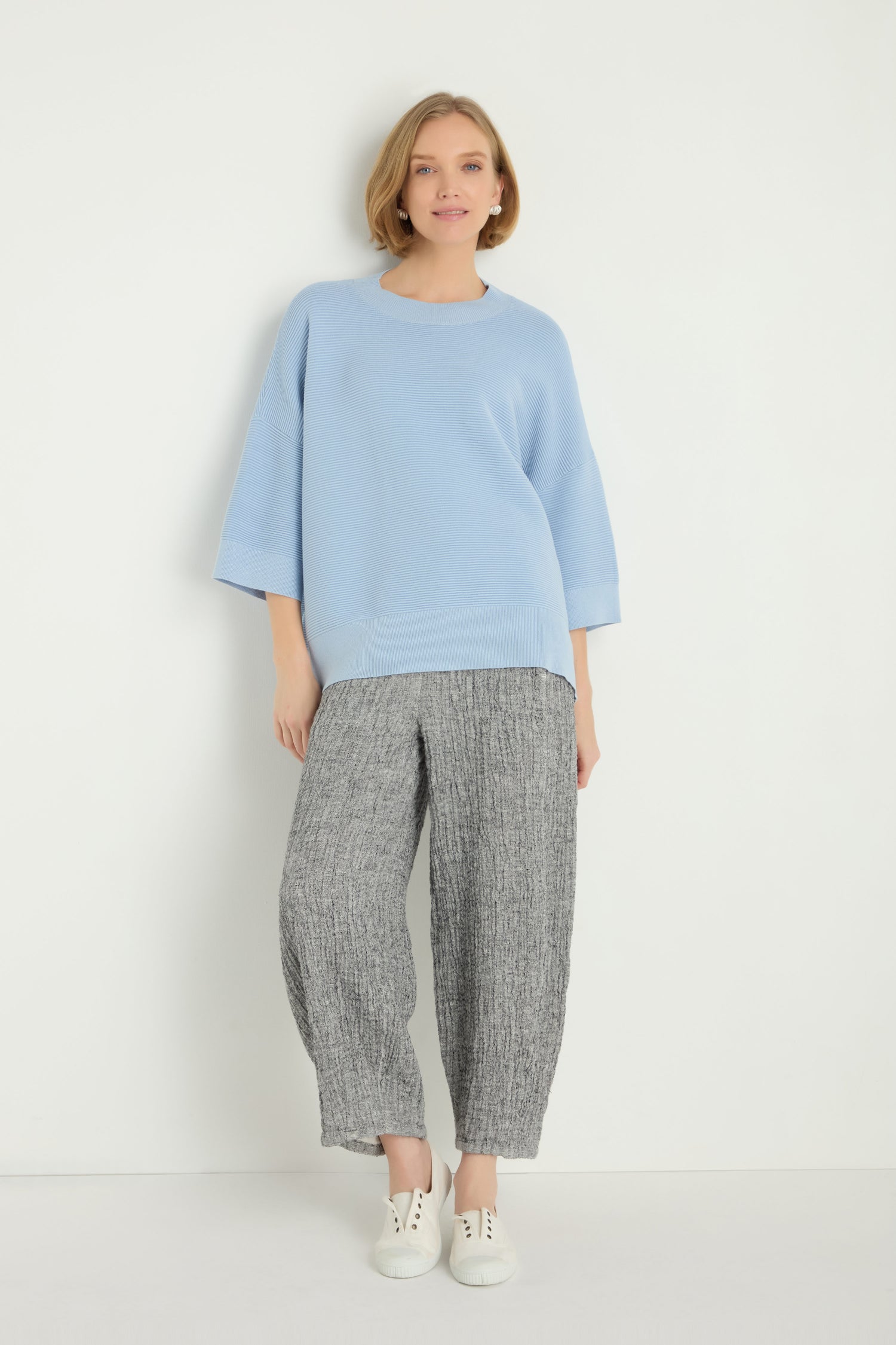 A woman poses by a white wall wearing the Wide Sleeve Ribbed Top in light blue, styled with gray wide-leg pants and white sneakers.