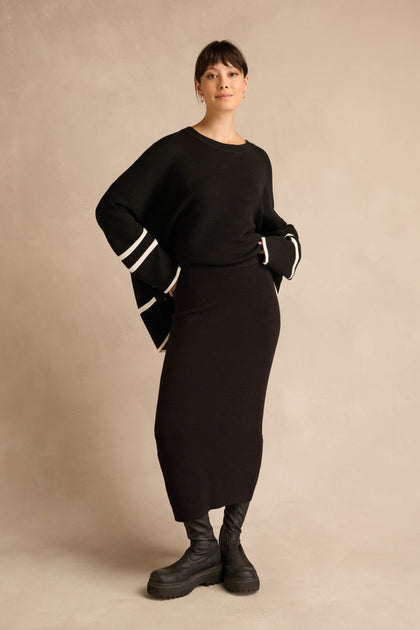 A woman stands against a beige background, wearing a black sweater with white stripes on the sleeves, the Ribbed Tube Skirt featuring a classic column silhouette that falls to midi length, and black boots.