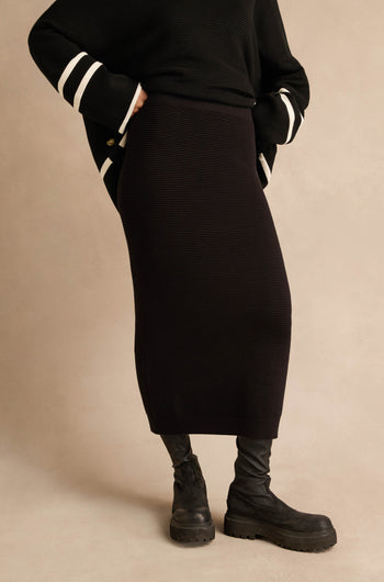 Wearing the Ribbed Tube Skirt in black—a classic, ribbed midi design—paired with a black sweater featuring white-striped sleeves, black tights, and chunky boots, this person stands against a neutral background.