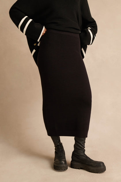 Wearing the Ribbed Tube Skirt in black—a classic, ribbed midi design—paired with a black sweater featuring white-striped sleeves, black tights, and chunky boots, this person stands against a neutral background.