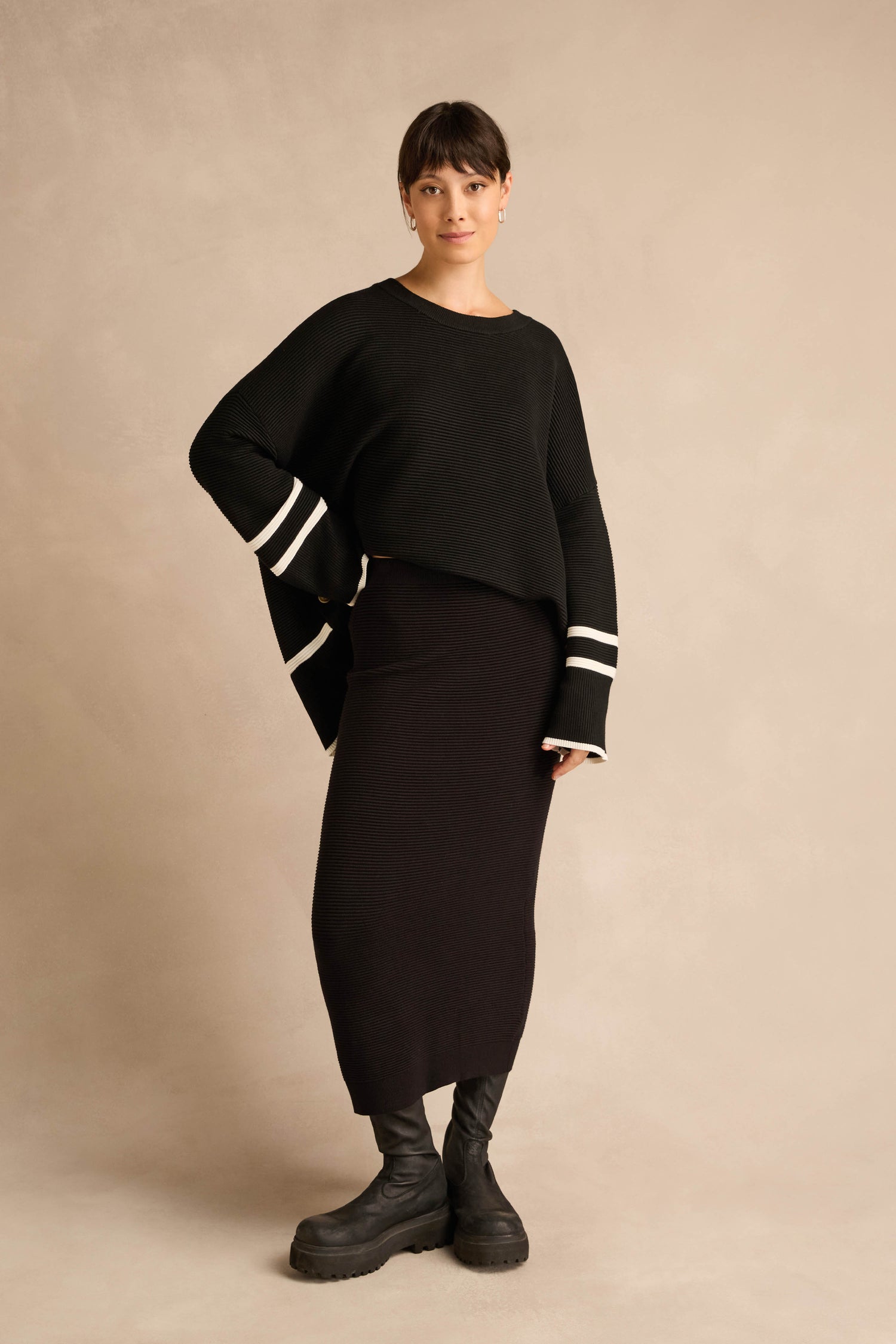 A person stands against a beige background, styled in a black oversized sweater with white sleeve stripes, the Ribbed Tube Skirt featuring a column silhouette and midi length, and black boots.