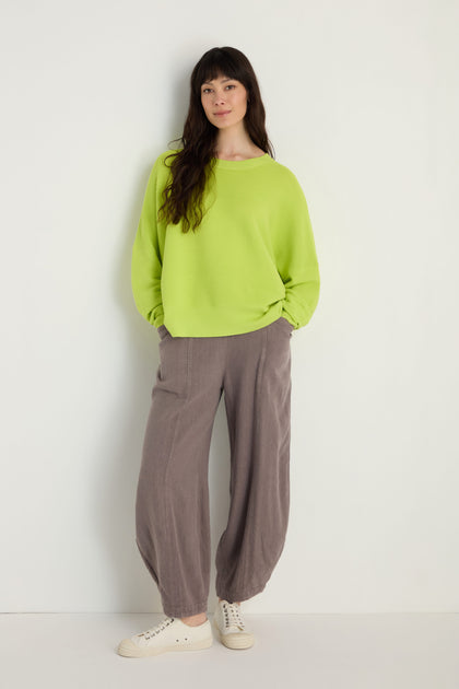 A woman stands against a plain wall, wearing Paisie's Boxy Ribbed Top in lime green with loose gray pants and white sneakers, her hands tucked casually in her pockets for a relaxed look.