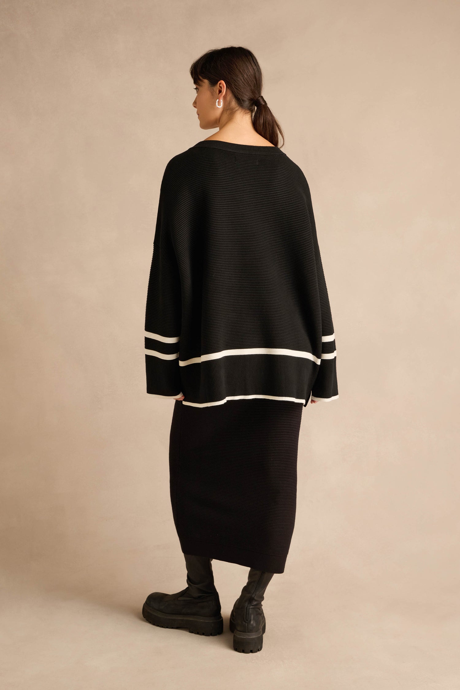 A woman stands facing away, wearing the Piping Detail Button Ribbed Top—an oversized black knit sweater with white stripes—paired with a black midi skirt, black boots, and hoop earrings against a beige background.