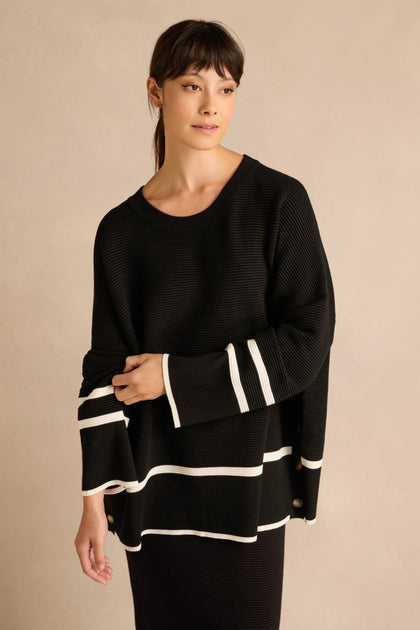 A woman wears the Piping Detail Button Ribbed Top, an oversized black knit sweater featuring white piping detail along the sleeves and hem, standing against a neutral beige background.