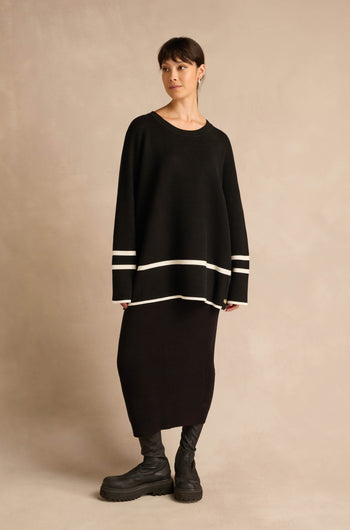 A woman stands against a beige background, wearing the Piping Detail Button Ribbed Top—an oversized black knit sweater featuring white striped piping on the sleeves and hem—paired with a long black skirt and black boots.