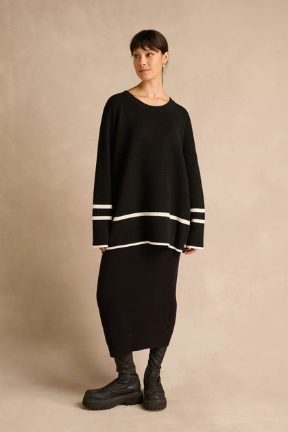 A woman stands against a beige background, wearing the Piping Detail Button Ribbed Top—an oversized black knit sweater featuring white striped piping on the sleeves and hem—paired with a long black skirt and black boots.