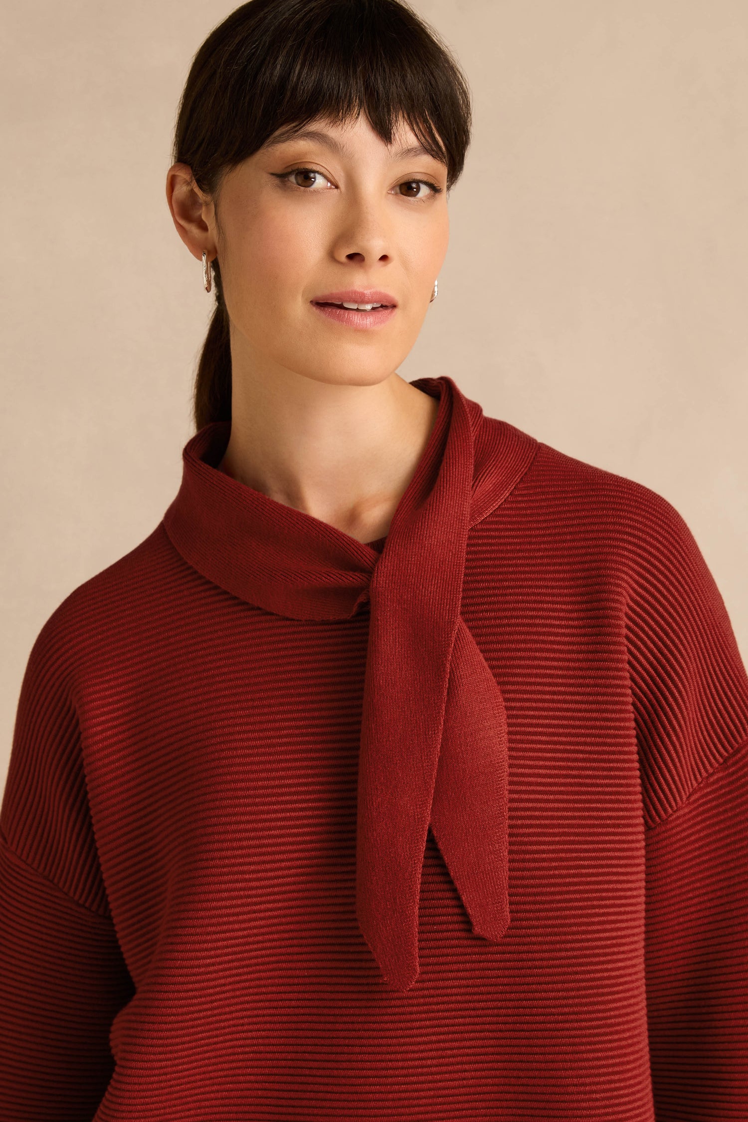 A woman with straight dark hair is wearing the Tie Neck Burgundy Ribbed Knit sweater, which features a tie neck detail and relaxed fit, standing against a neutral beige background.