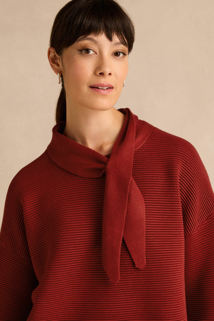A woman with straight dark hair is wearing the Tie Neck Burgundy Ribbed Knit sweater, which features a tie neck detail and relaxed fit, standing against a neutral beige background.