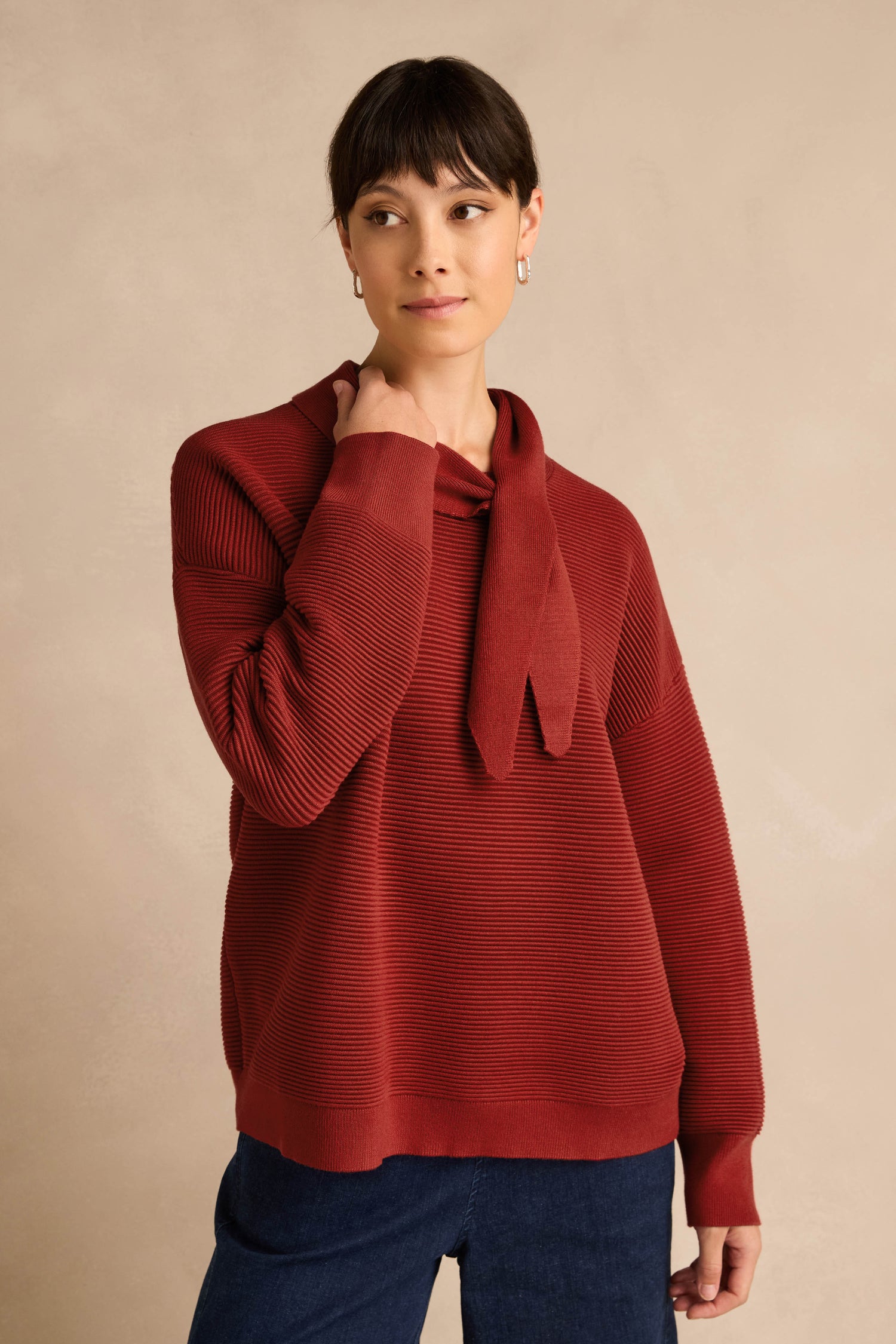 Wearing the Tie Neck Burgundy Ribbed Knit in a relaxed fit, a person stands against a beige background, gazing to the side with one hand touching their neck.