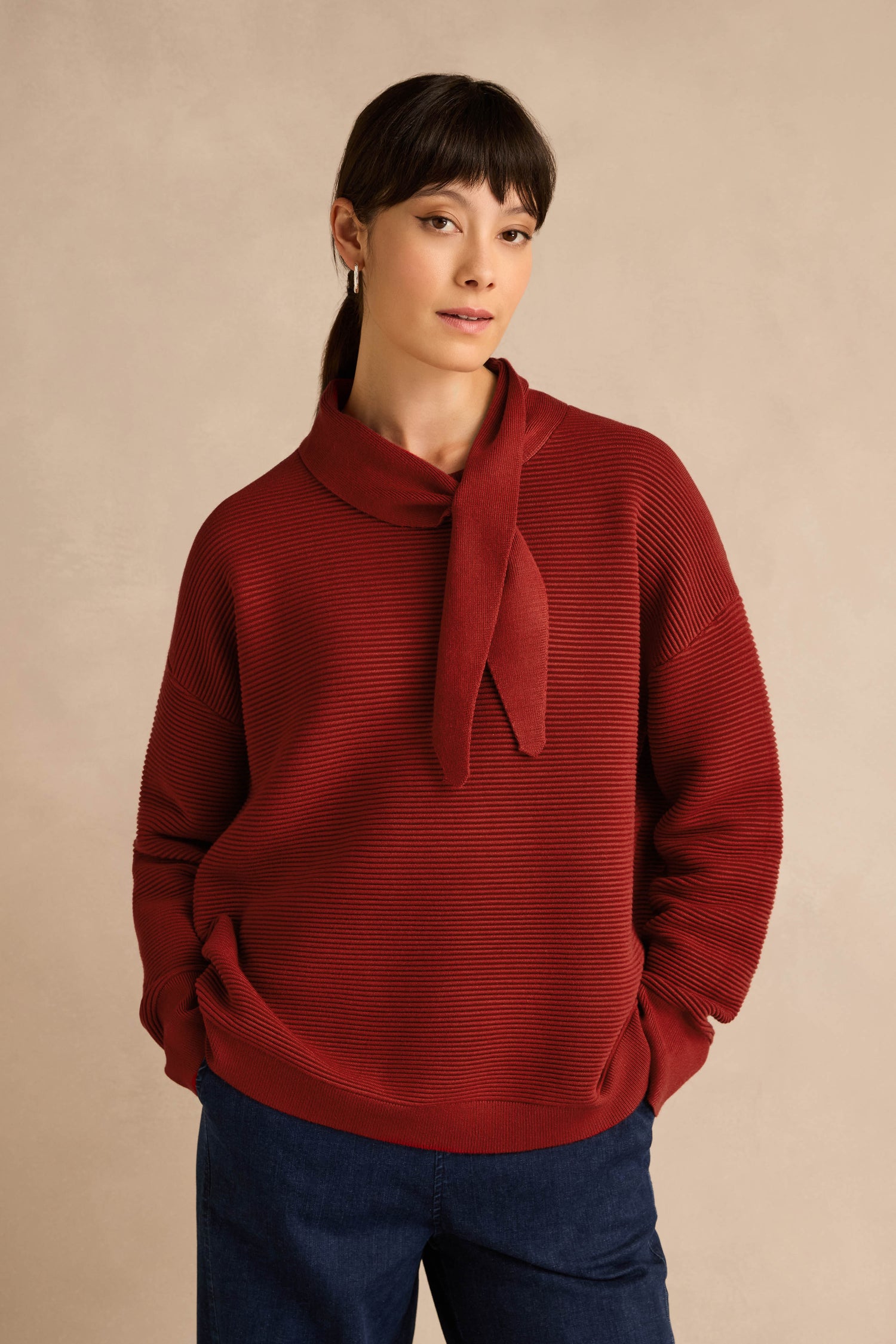 A woman stands against a beige background wearing the Tie Neck Burgundy Ribbed Knit sweater, styled in a relaxed fit with a ribbed knit collar and paired with dark blue pants. Her hands are casually in her pockets as she looks at the camera.
