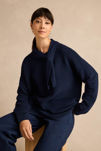 A woman is seated on a stool, wearing the Tie Neck Navy Ribbed Knit sweater and dark blue jeans, set against a plain beige background.