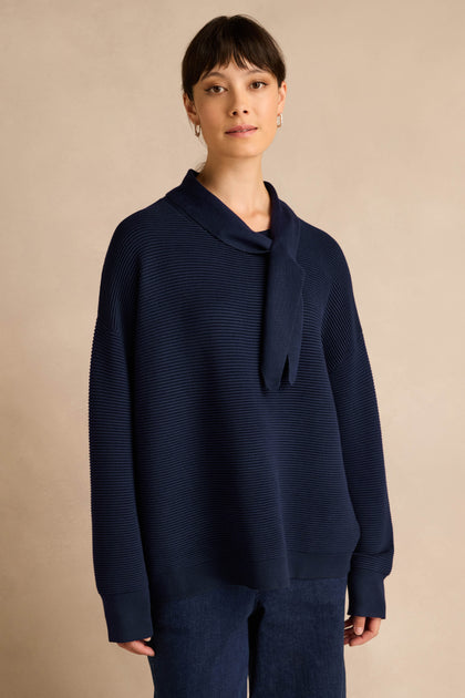 A woman stands against a beige background, wearing the Tie Neck Navy Ribbed Knit sweater paired with dark pants. She looks directly at the camera with a neutral expression, while the relaxed fit and collar detail of the jumper add effortless sophistication to her look.