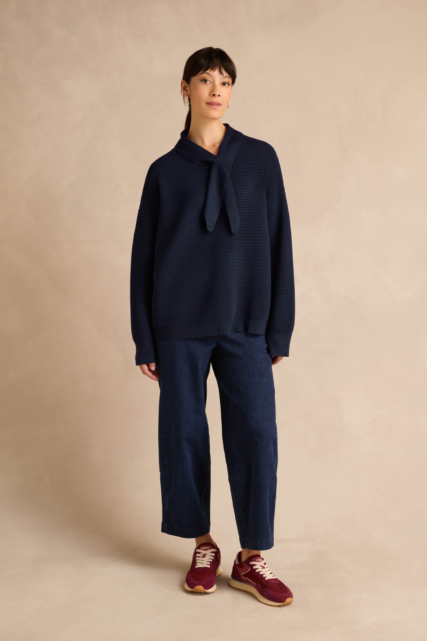 A woman stands against a beige textured background wearing the Tie Neck Navy Ribbed Knit jumper with a relaxed fit, paired with wide-leg dark blue pants and red sneakers.