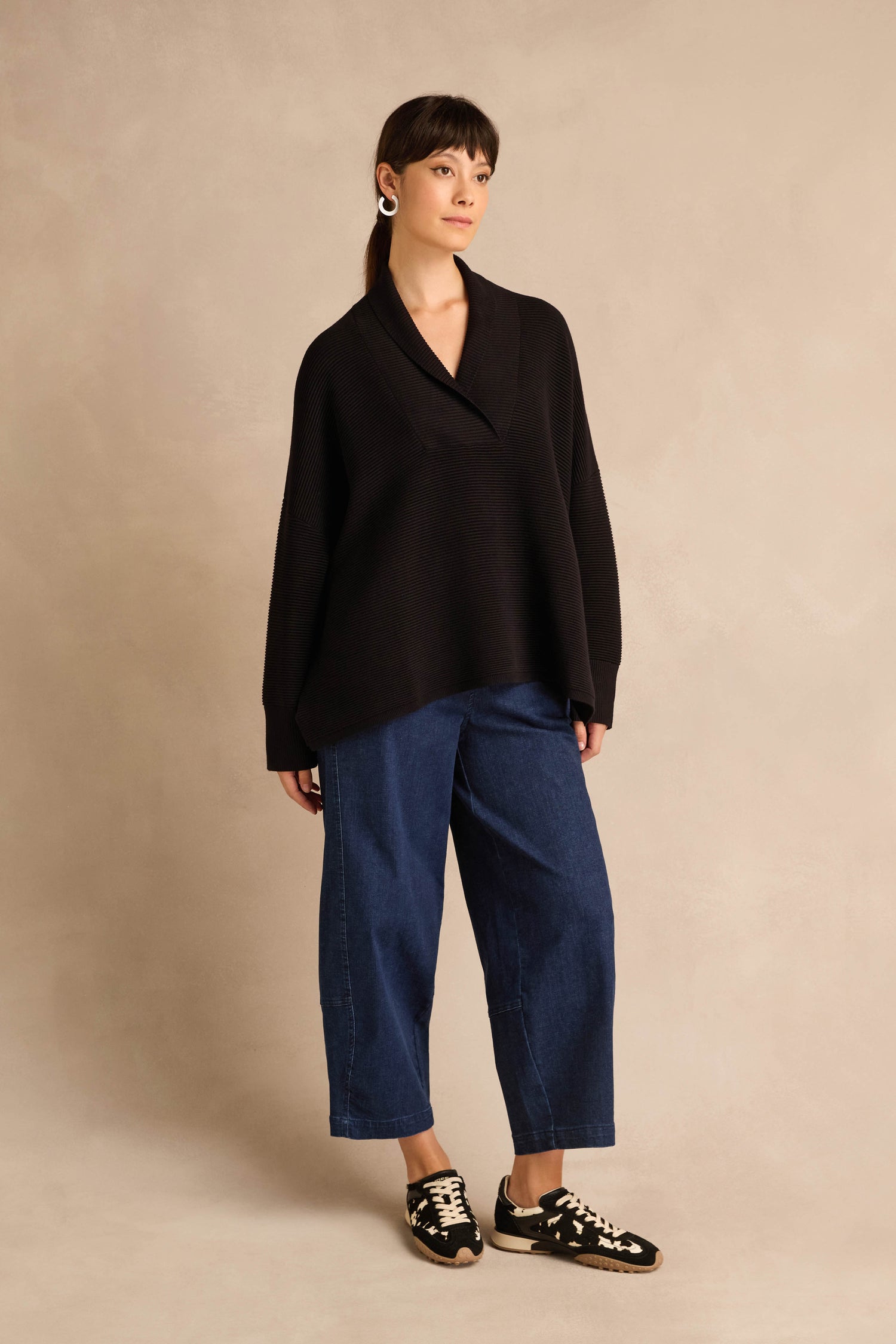 Woman stands against a beige background wearing a relaxed fit, loose Shawl Collar Black Ribbed Knit sweater, wide-leg blue jeans, black sneakers, and silver earrings.