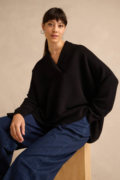 A woman with straight dark hair and hoop earrings is seated on a wooden stool against a beige background, wearing the Shawl Collar Black Ribbed Knit sweater with a relaxed fit and blue jeans.