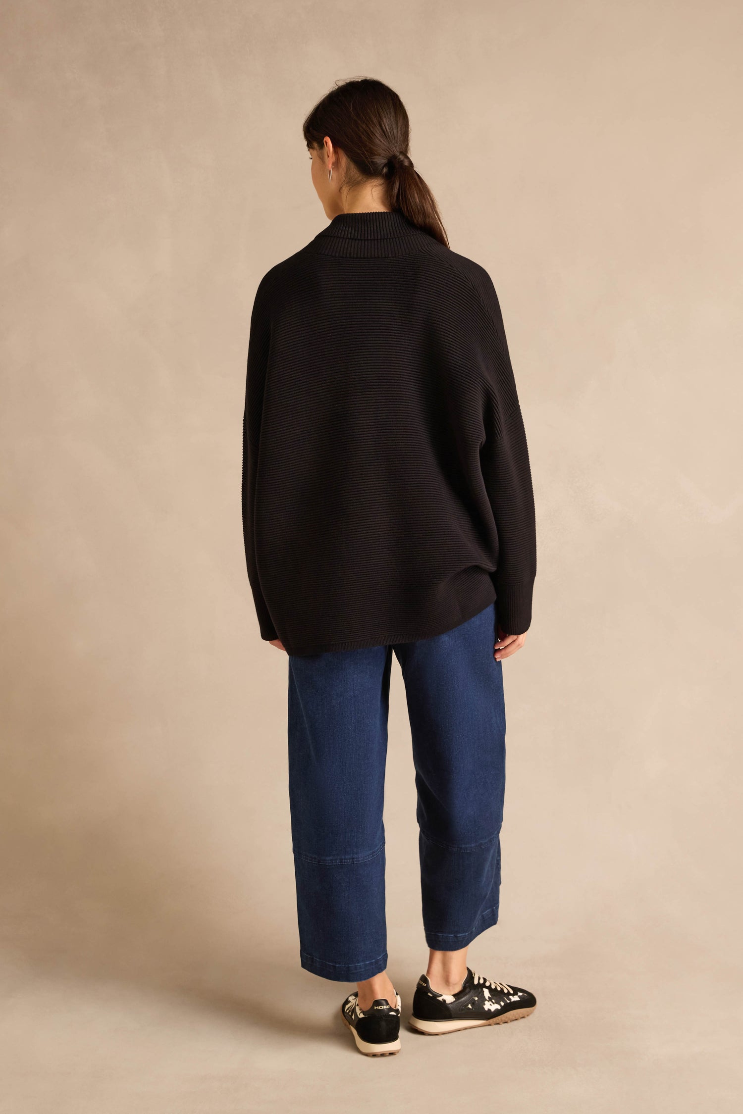Person standing with back to camera, wearing the Shawl Collar Black Ribbed Knit sweater, blue wide-leg jeans, and black sneakers, against a light beige background.