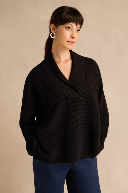 A woman stands against a beige background, wearing the Shawl Collar Black Ribbed Knit sweater in a relaxed fit with blue jeans and hoop earrings, looking to the side with her hands in her pockets.
