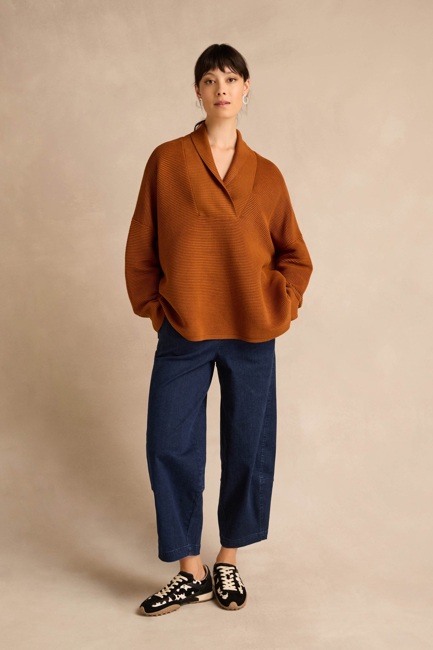 A woman stands against a beige background wearing the Shawl Collar Copper Ribbed Knit pullover, blue wide-leg jeans, and black-and-white sneakers.