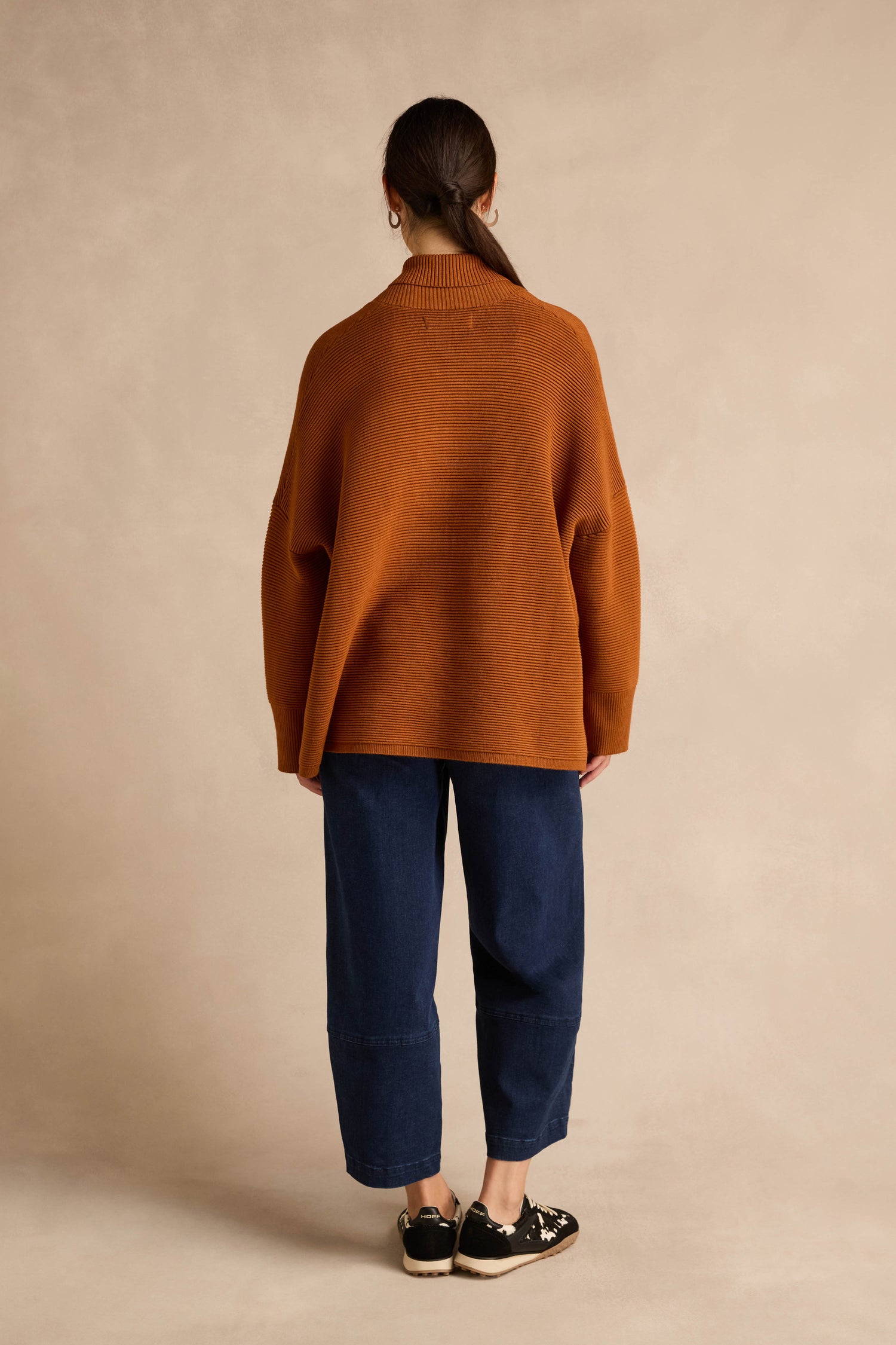 A person with long brown hair, wearing the Shawl Collar Copper Ribbed Knit sweater with a relaxed fit, dark blue pants, and black sneakers stands facing away from the camera against a beige background.