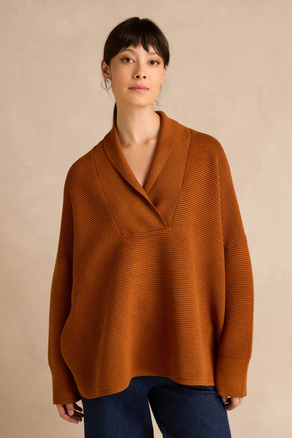 A woman wearing the Shawl Collar Copper Ribbed Knit—a loose, relaxed fit sweater—with dark blue jeans stands in front of a beige background.