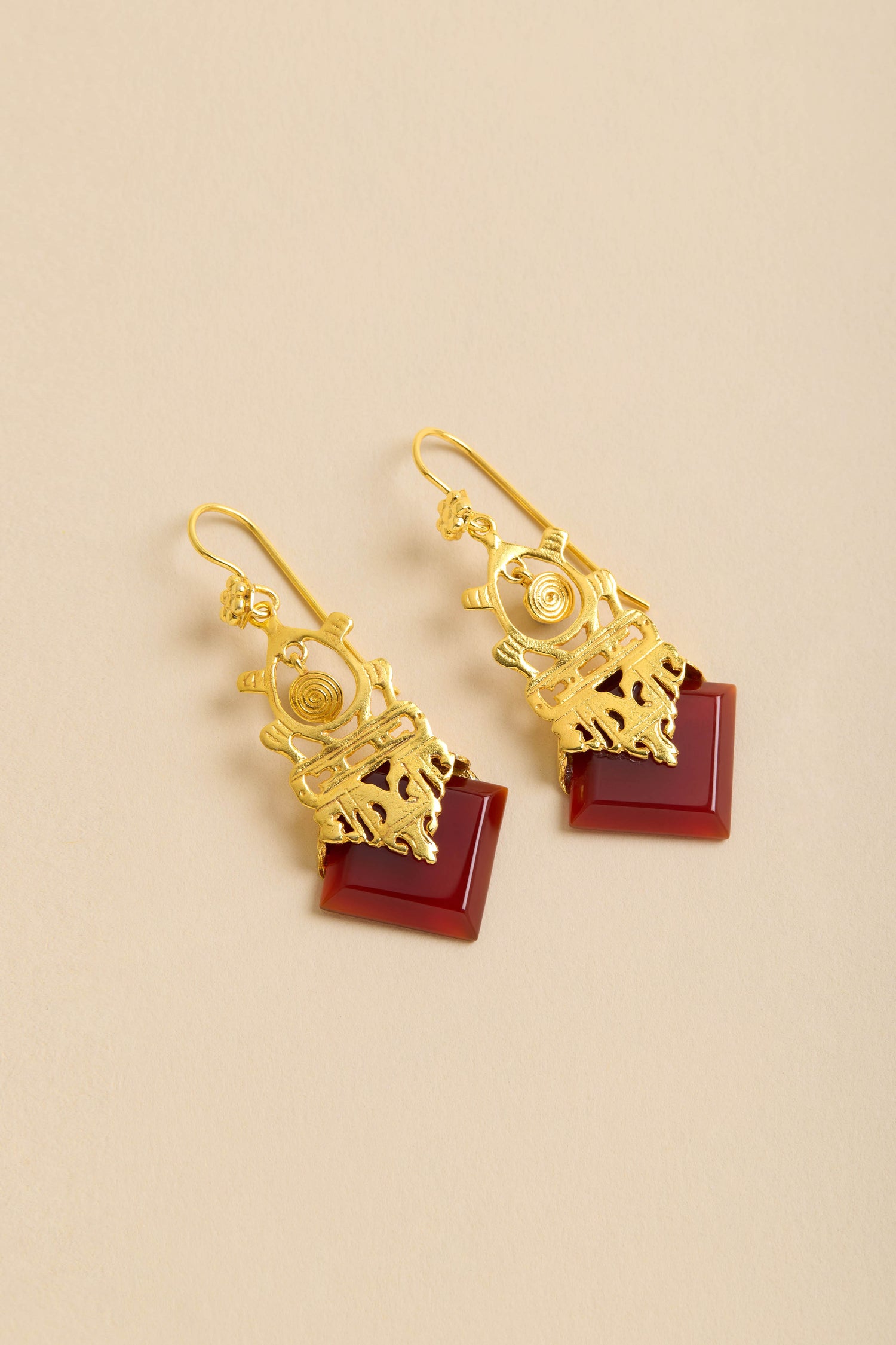 Gold-Plated Carnelian Drop Earrings featuring intricate designs and square reddish-brown stone accents, shown on a beige background.