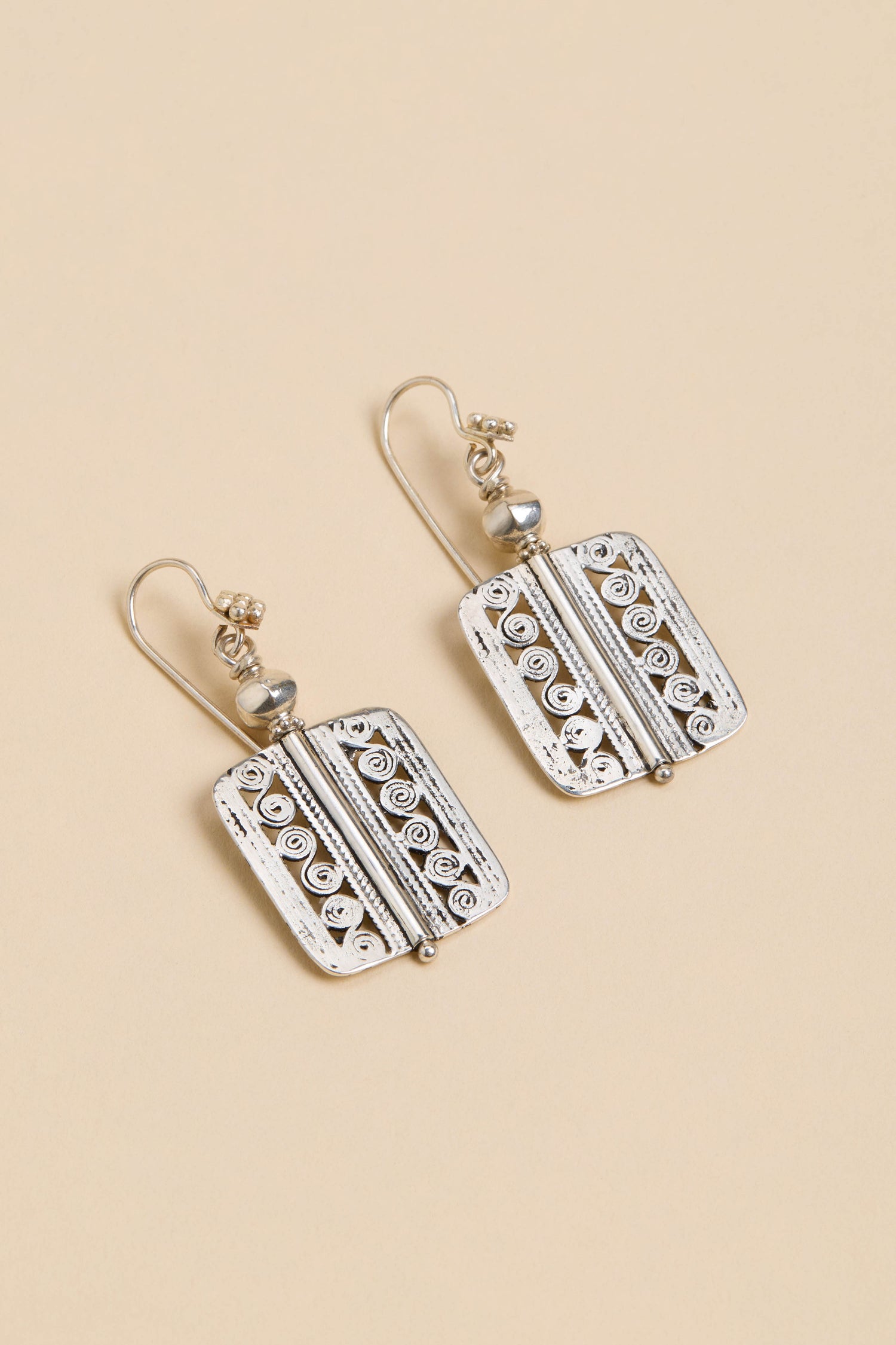 Filigree Drop Earrings—rectangular silver drops with swirl and line details, small round accents, and hooks, shown on a beige background.