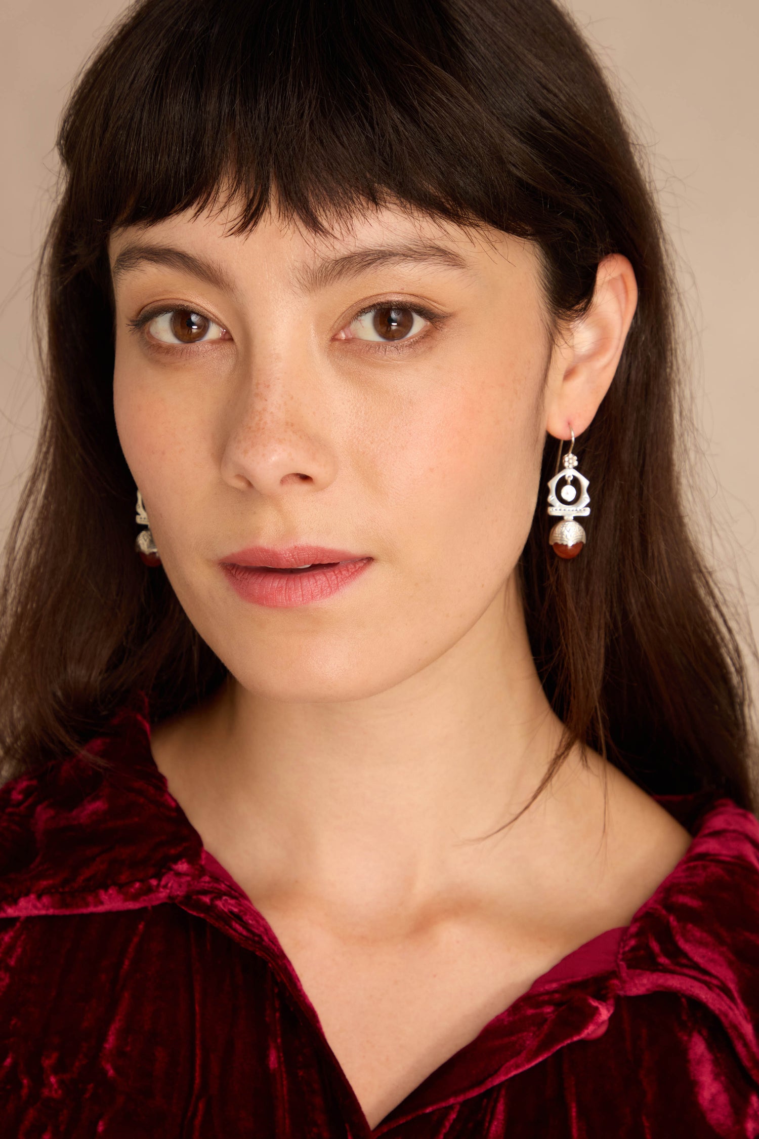 Woman with straight brown hair, in a red velvet top and Carnelian Drop Earrings, looks at the camera against a light beige background.