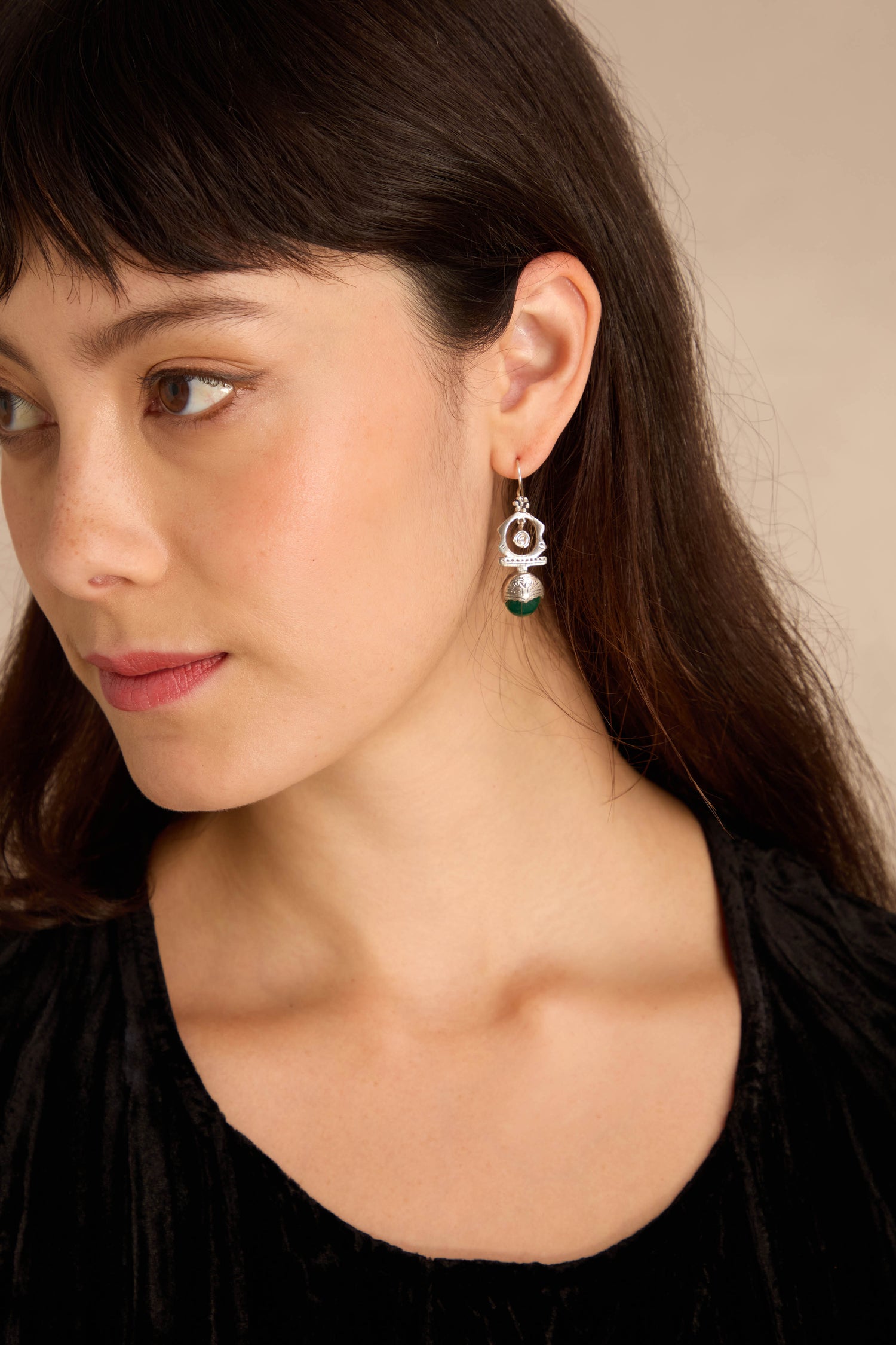 A woman with long dark hair, in a black top, wears Green Onyx Drop Earrings and looks left against a plain beige background.