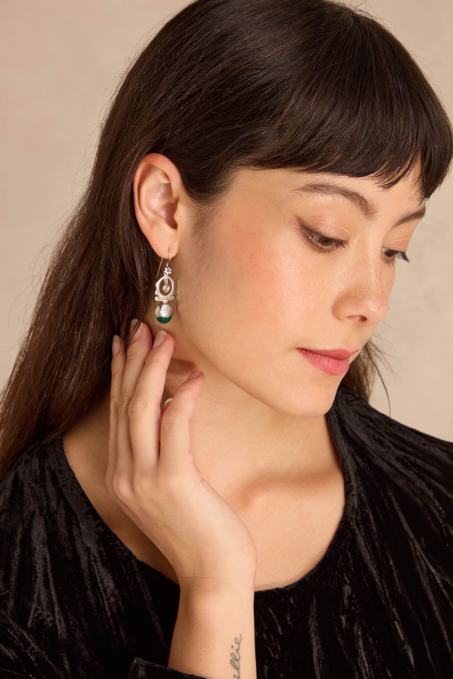 A woman with long dark hair touches her Green Onyx Drop Earrings set in sterling silver. She has natural nails and a small tattoo on her wrist.