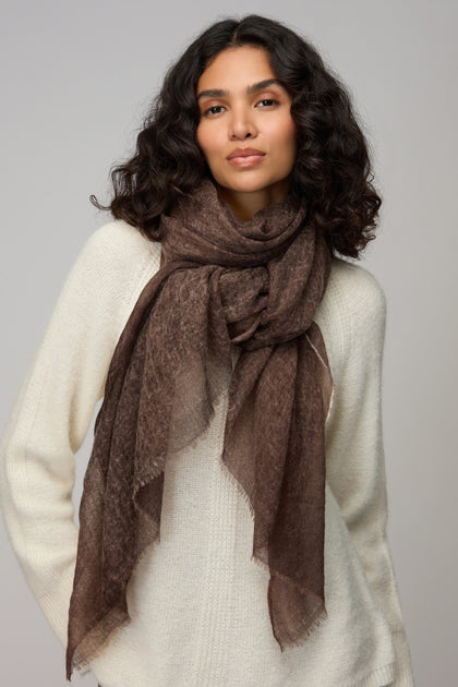 A woman with wavy dark hair wears a white sweater and the Soft Espresso Cashmere Scarf, standing against a plain light gray background.