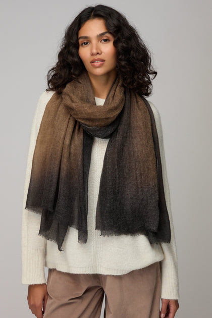 A woman in a cream sweater and light brown pants stands against a plain light background, accessorized with the Espresso Gradient Cashmere Scarf—a luxurious brown-to-black gradient piece.