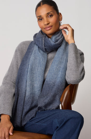 A woman seated on a chair wears a grey sweater, blue jeans, and the Night Sky Gradient Cashmere Scarf, with one hand touching her earring.