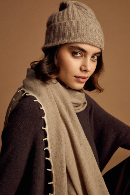 A woman models the Warm Sands Ribbed Cashmere Hat with a matching scarf and dark sweater against a plain brown background—a stylish winter accessory look.