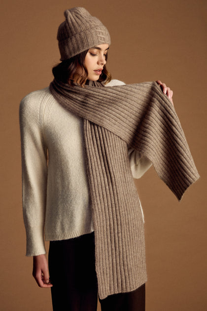 A woman wearing a cream sweater, brown knit hat, and the Warm Sands Ribbed Cashmere Scarf stands against a brown background—an ideal touch to enhance your winter wardrobe.