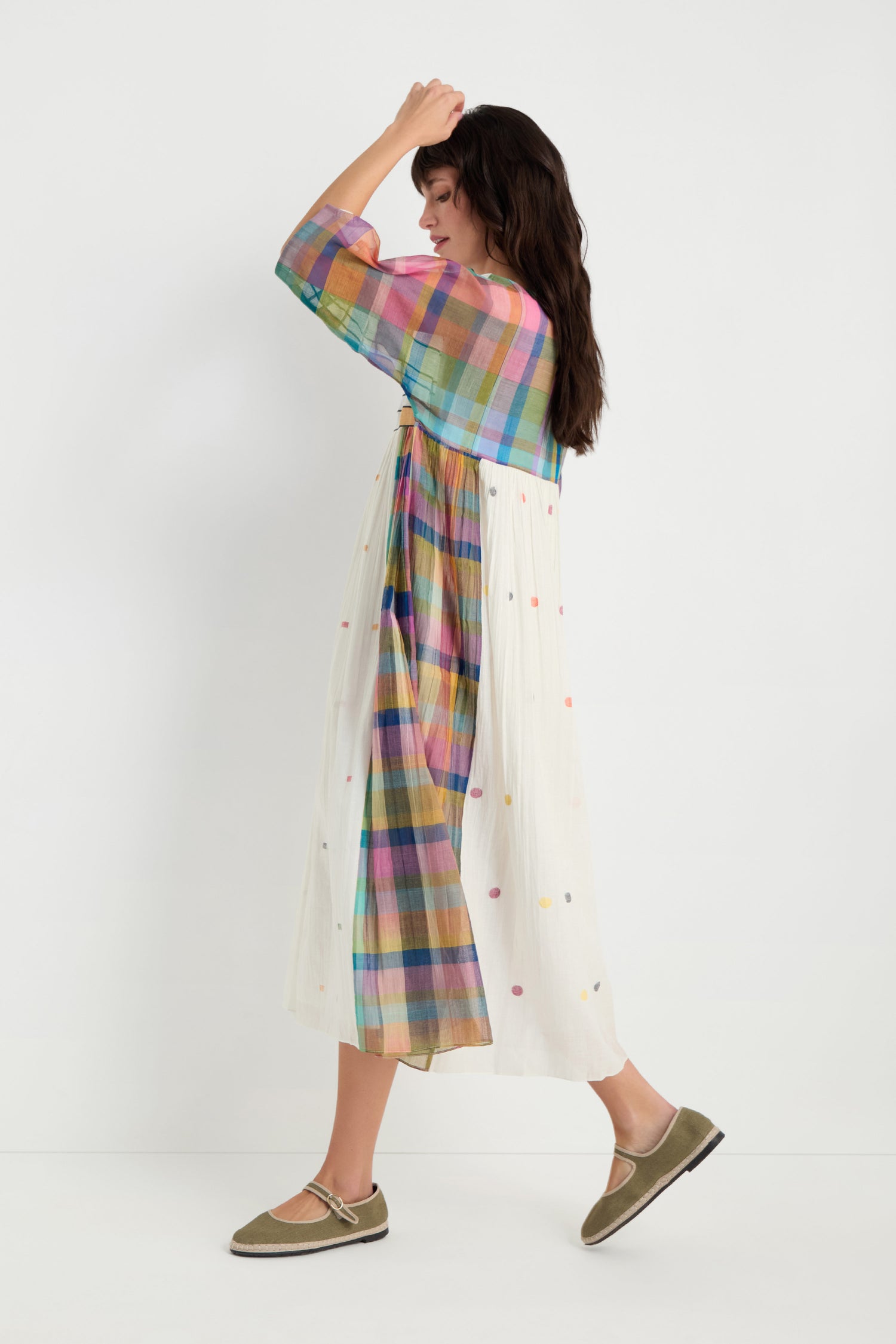 A woman in profile models the Mondrian Jamdani Dress featuring a colorful plaid top and white polka-dot skirt, paired with green flats, against a plain white background.