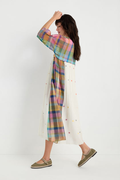 A woman in profile models the Mondrian Jamdani Dress featuring a colorful plaid top and white polka-dot skirt, paired with green flats, against a plain white background.