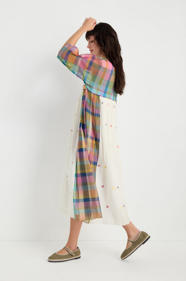 A woman in profile models the Mondrian Jamdani Dress featuring a colorful plaid top and white polka-dot skirt, paired with green flats, against a plain white background.
