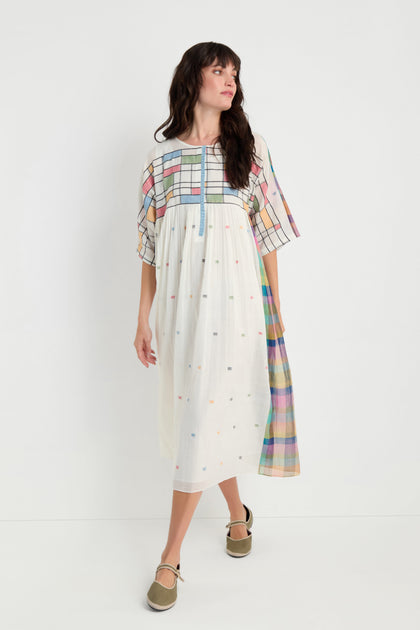 A woman models the Mondrian Jamdani Dress—a loose, knee-length cotton dress with pastel plaid and polka dot patterns—standing against a plain white background.