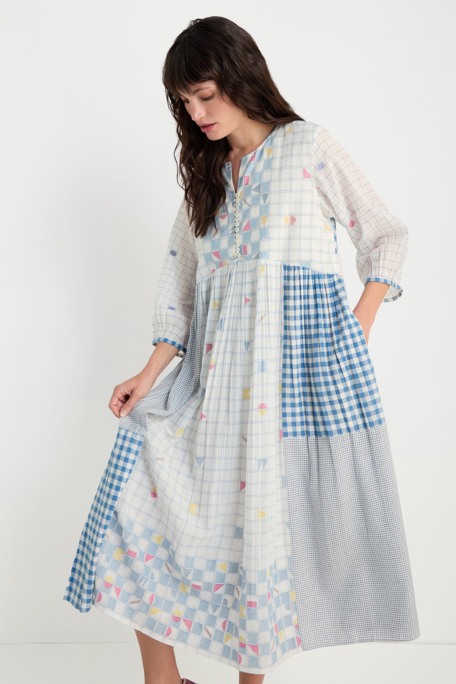 A woman stands against a plain white background wearing the Jamdani Check Dress—a long-sleeved, pure cotton midi with blue, white, and pastel check patterns.
