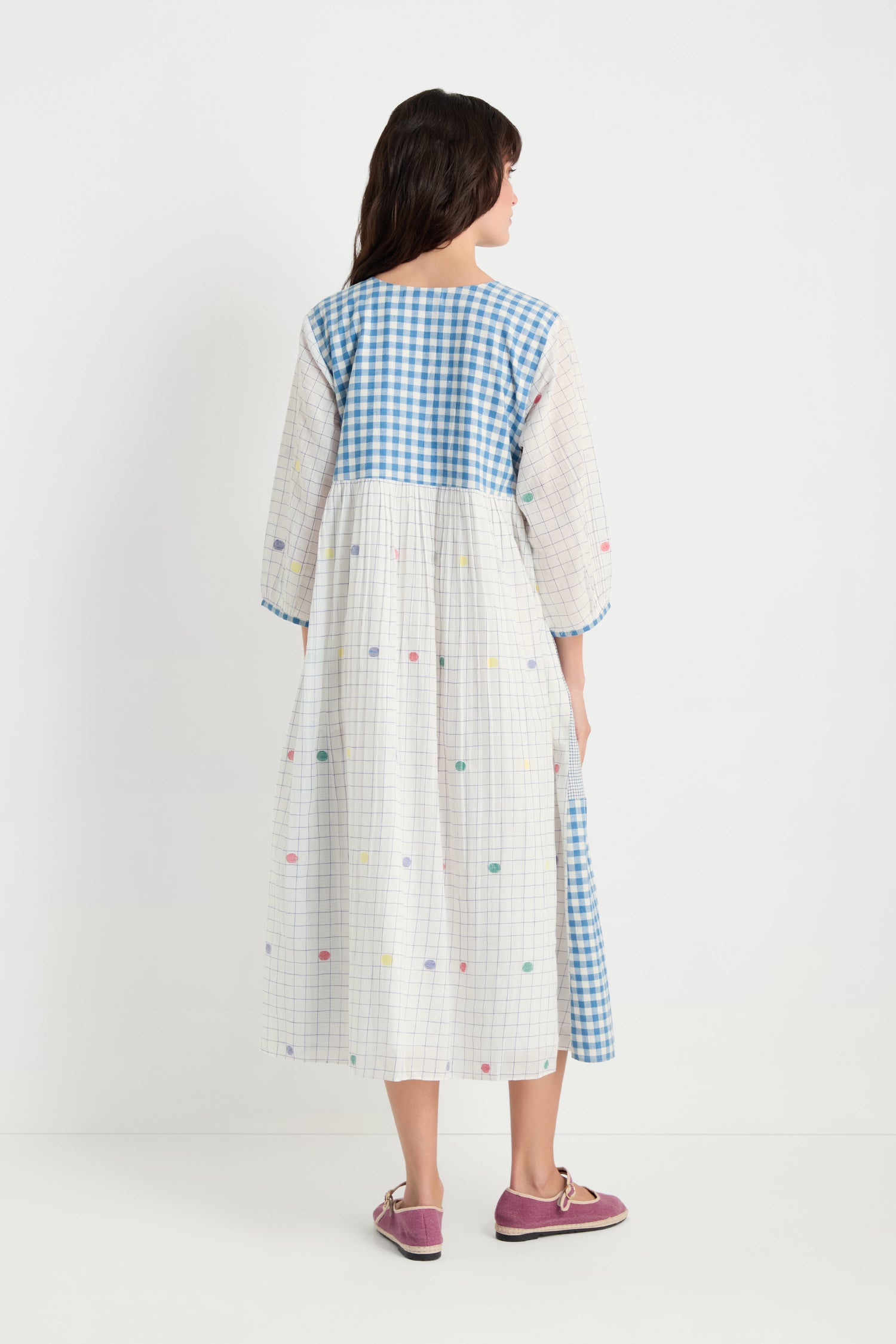 A woman faces away, wearing the Jamdani Check Dress—a pure cotton midi dress with a blue gingham yoke and multicolored dots—paired with pink shoes.