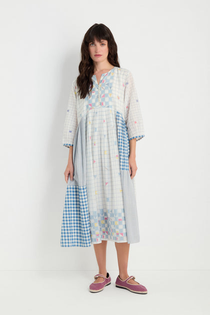 A woman stands against a white background wearing the Jamdani Check Dress, made of pure cotton with mixed pastel checks in a midi length, paired with lavender shoes.