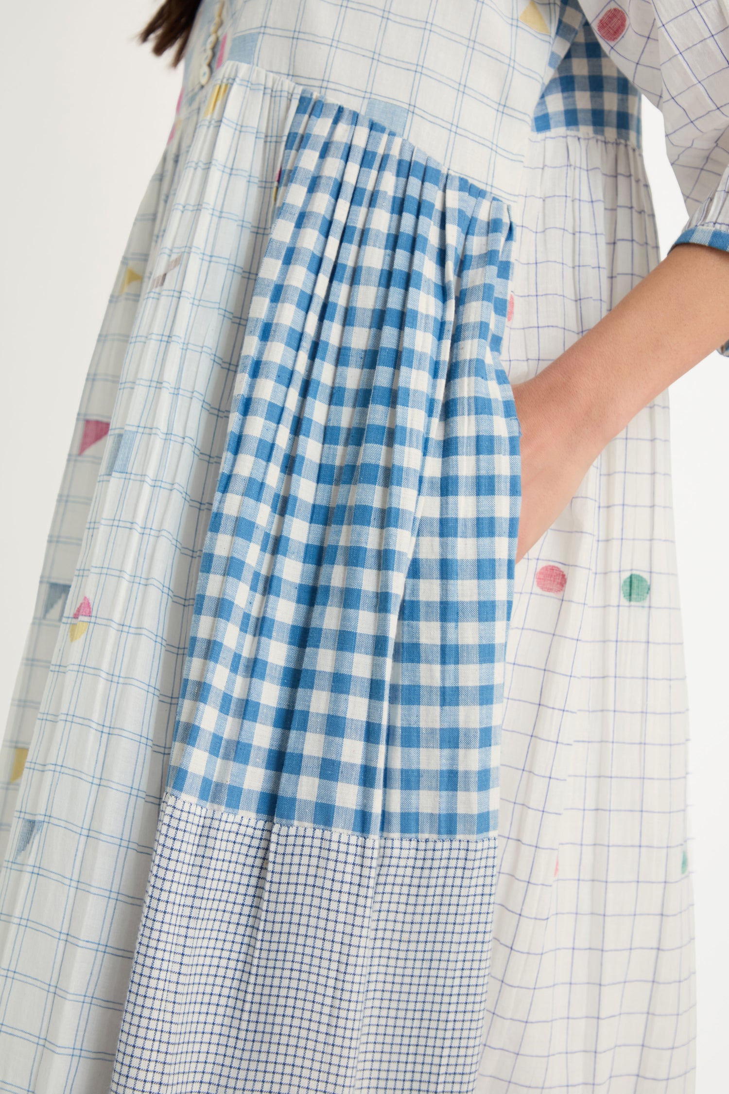 A person wearing the Jamdani Check Dress, a pure cotton midi dress with blue and white check patterns and a grid print, stands with one hand in the pocket.