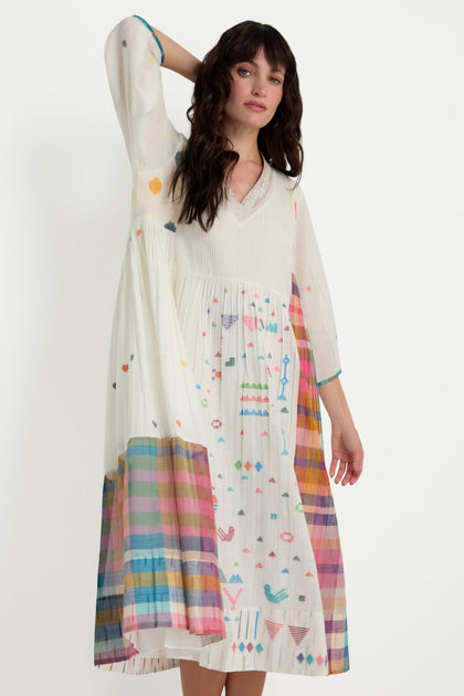 A woman models the Jamdani Check Pleated Dress—long, white cotton with colorful geometric patterns and a striped hem—posing with one hand raised to her head against a plain background.