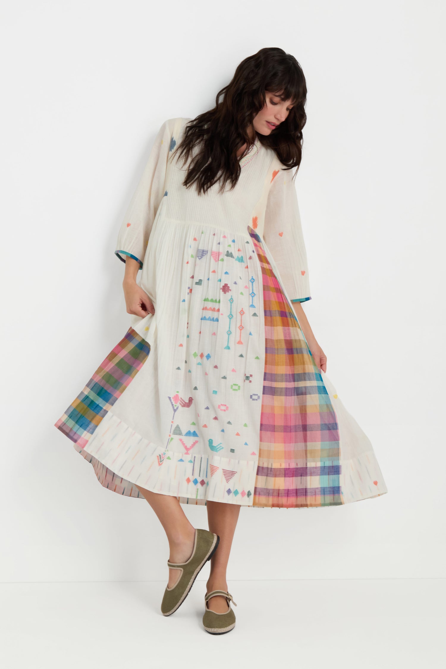 A woman wearing the Jamdani Check Pleated Dress—long, white cotton with colorful embroidery and a rainbow plaid panel—stands against a plain white background.