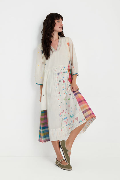A woman models the Jamdani Check Pleated Dress, featuring vibrant patterns and multicolored pleated panels, as she stands against a plain white background looking to the side.
