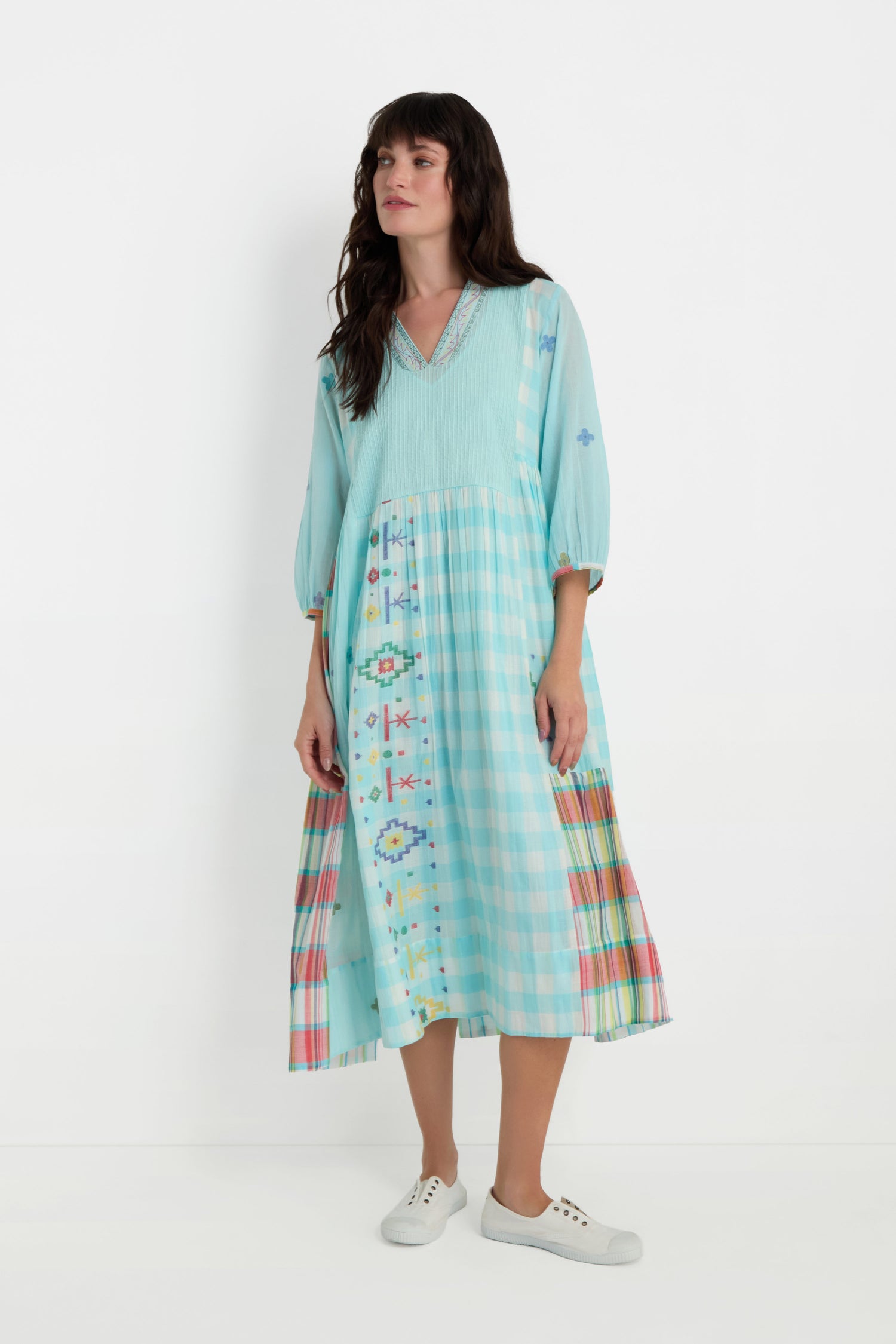 Woman wears the Pintuck Jamdani Dress—a light blue, loose cotton dress with checkered and colorful woven motifs, pintuck front panel, and three-quarter sleeves—with white sneakers, standing against a plain white background.